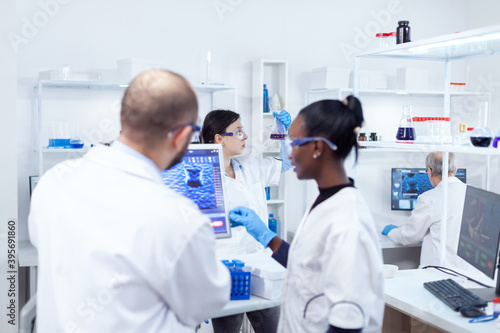 Scientist holding glass flask with blue genetic material in busy laboratory. Multiethnic team of medical researchers working together in sterile lab wearing protection glasses and gloves.