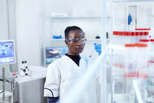 African scientist in protective glasses looking at test tube with genetic material. Black researcher in sterile laboratory conducting pharmacology experiment wearing coat.