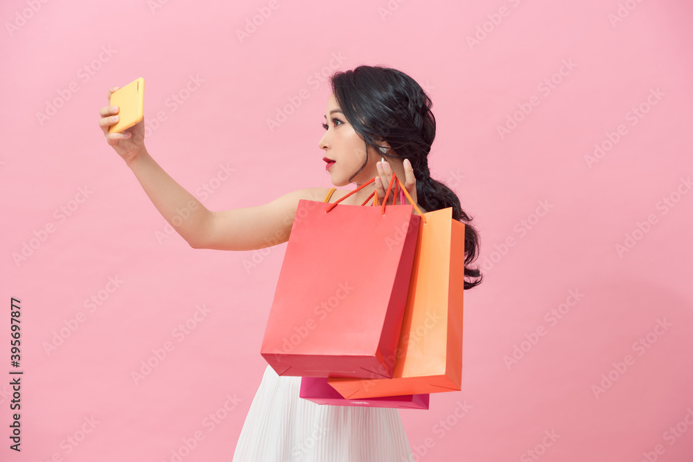 Fototapeta premium Happy pretty young woman with shopping bags makes photo self-portrait on the smartphone over colorful wall