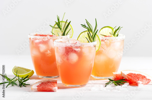 Three glasses of paloma cocktail with ice and tequila, decorated with lime wedges, grapefruit and rosemary stand on a white wooden table. Horizontal orientation, selective focus
