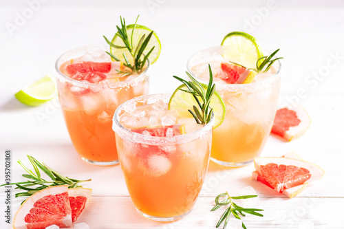 Paloma cocktail with ice and tequila, garnished with lime wedges, grapefruit and rosemary on a white wooden table. Horizontal orientation, selective focus