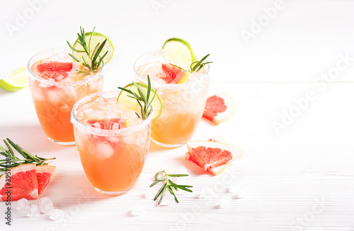 Three paloma alcoholic cocktails with ice and tequila, garnished with lime wedges, grapefruit and rosemary stand on a white wooden table. Horizontal orientation, selective focus, copy space.