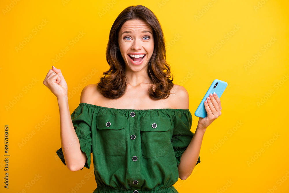 Photo of brown curly haired young attractive woman raise fist lucky winner hold phone isolated on shine yellow color background Photo of brown curly haired young attractive woman raise fist lucky winner hold phone isolated on shine yellow color background