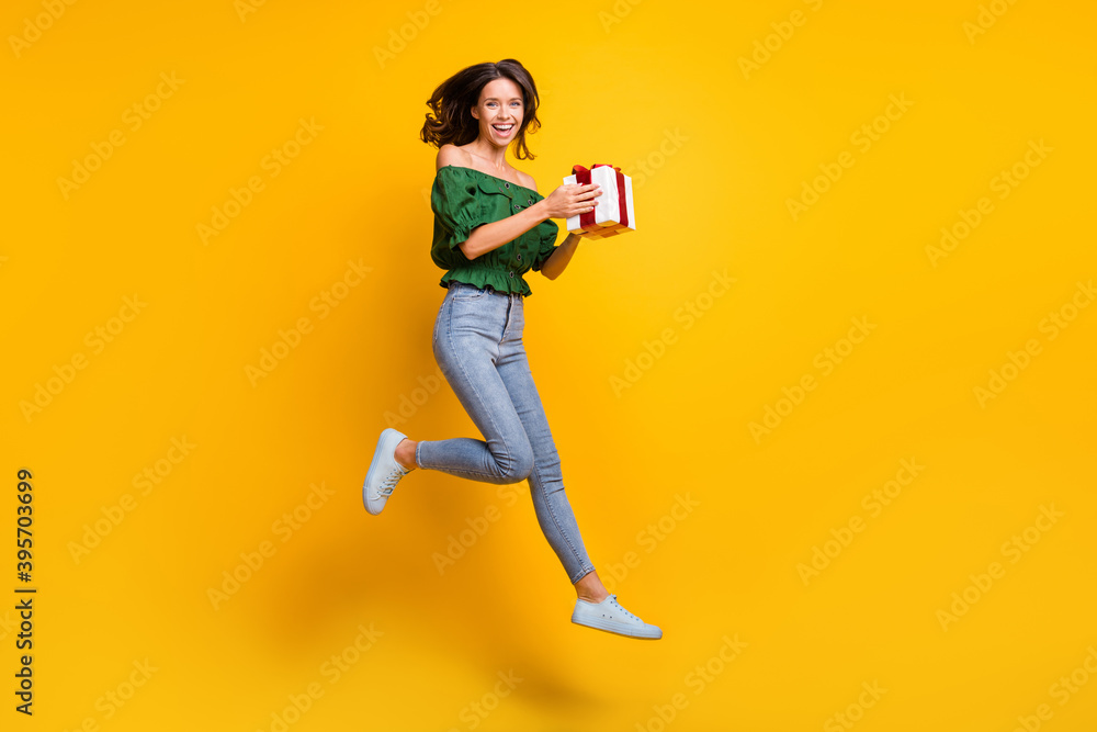 Full size profile side photo of attractive nice young lady jump up air hold gift box wear jeans isolated on shine color background Full size profile side photo of attractive nice young lady jump up air hold gift box wear jeans isolated on shine color background
