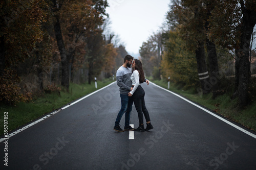 Beautiful portrait of young couple hugging and standing on a perfect clean long road bewtween the green forest in autumn