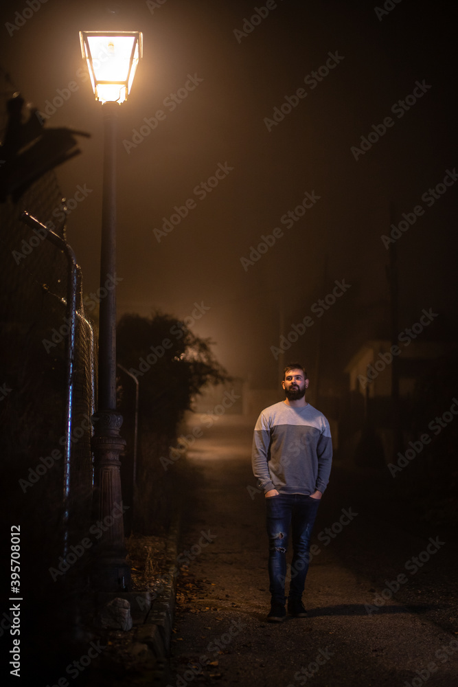 Young handsome man with black beard walking under the light of a street light at night time
