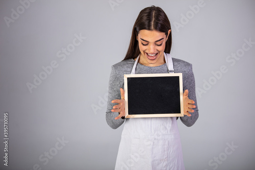 emale Employee Wearing Uniform Apron Holding Blackboard Sign. Friendly smiling shop assistant holding blank chalkboard sign isolated on gray background.