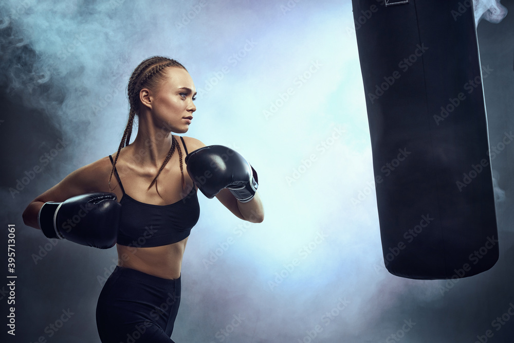 sporty brunette woman in boxing gloves and sportswear kicking bag on dark background with smoke.