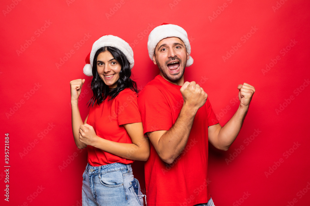 Fototapeta premium Cheerful young couple with win gesture in Santa Claus hats together isolated over red background