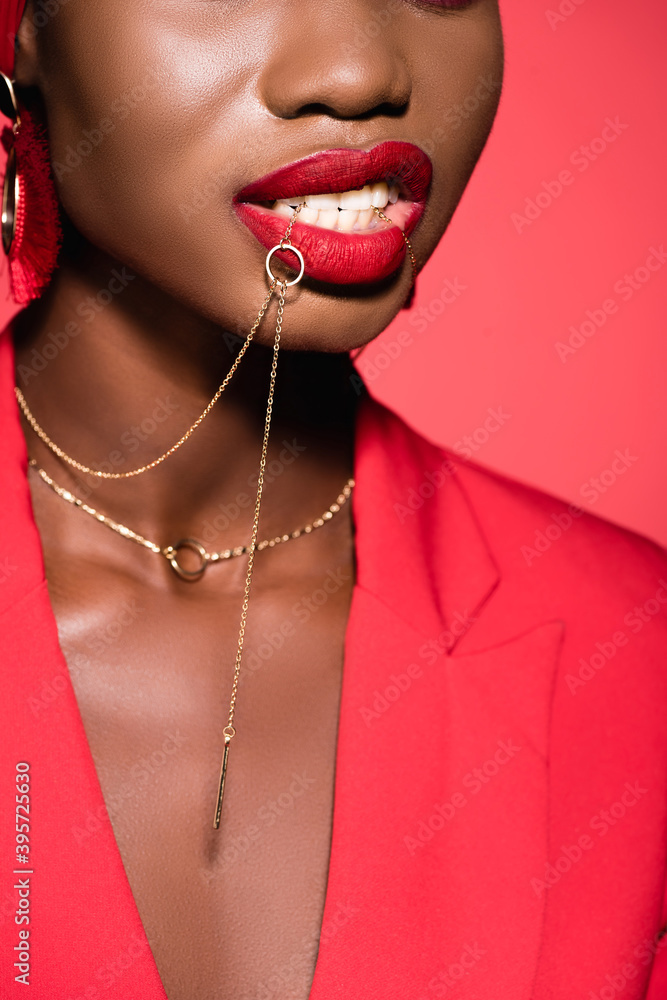 cropped view of african american woman in stylish outfit holding necklace in mouth isolated on red