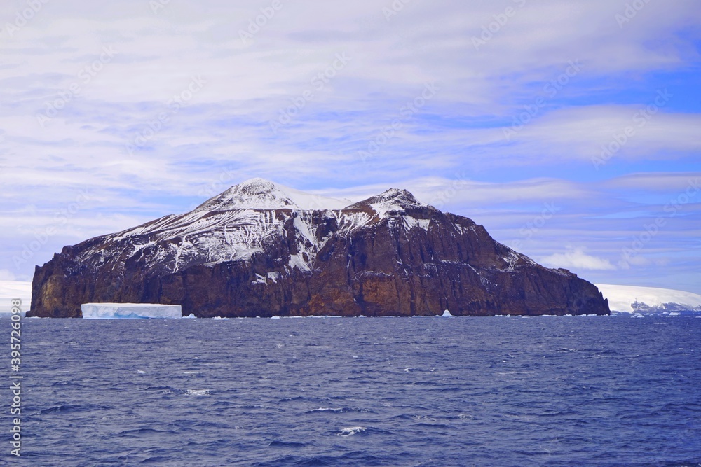 There is a rocky mountain in the sea, the top of which is covered with some snow. There is a small iceberg in front of it.