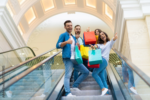 Four stylish smiling excited beautiful modern friends in casual clothes with paper bags are having fun while standing on the escalator in the mall during shopping.