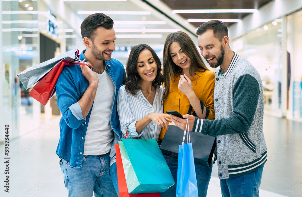 Obraz premium Stylish happy excited friends with shopping bags are looking together on the smart phone and chatting about sales in the mall