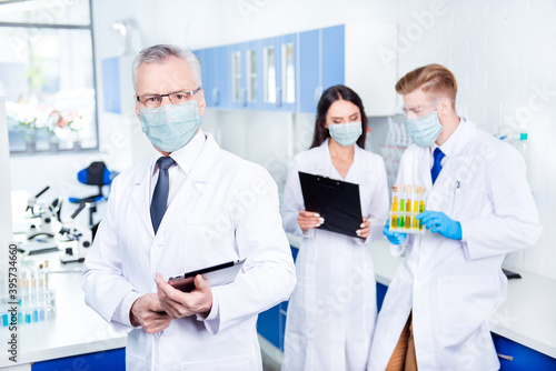 Portrait of professor in lab coat and glasses holding tablet making examination vaccine teamwork wearing gauze mask
