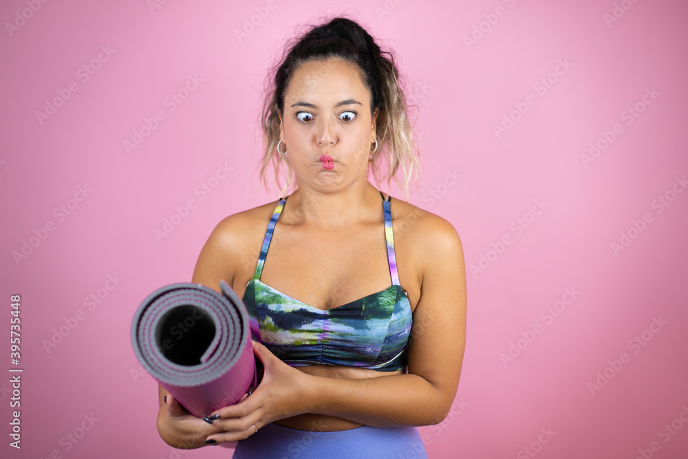 Young beautiful woman wearing sportswear and holding a splinter over isolated pink background making fish face with lips, crazy and comical gesture. Young beautiful woman wearing sportswear and holding a splinter over isolated pink background making fish face with lips, crazy and comical gesture.