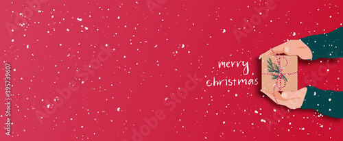 Female's hands in pullover holding Christmas gift box decorated with evergreen branch on red background with snow. Christmas and New Year banner.