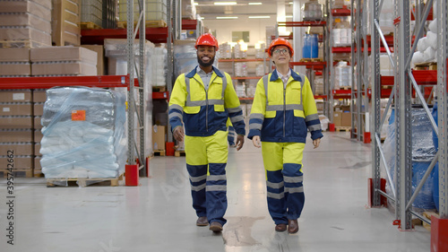 Two young engineers in workwear and hardhat walking along large warehouse photo