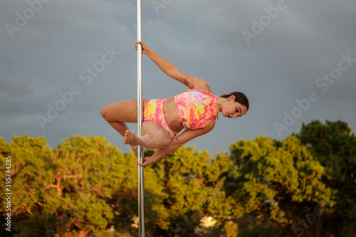 Young Middle Eastern pole dancer, holding a pose on a pole set in an urban park.