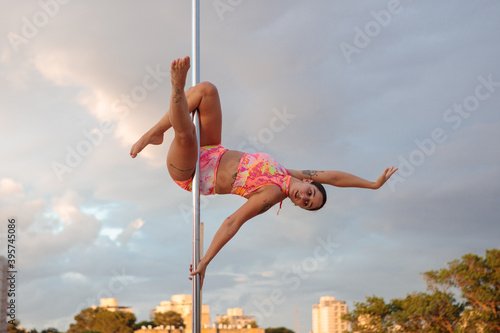 Young Middle Eastern pole dancer, holding a pose on a pole set outdoors. Tel Aviv buildings in the far background.