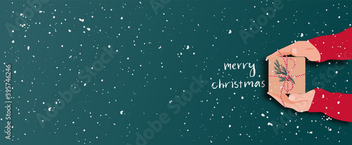 Female's hands in pullover holding Christmas gift box decorated with evergreen branch on red background with snow. Christmas and New Year banner.