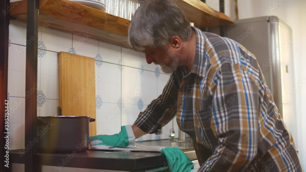 Senior man in gloves cleaning electric stove panel in kitchen