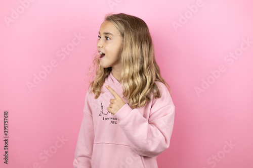 Young beautiful child girl standing over isolated pink background pointing with hand and finger to the side with surprised expression © Irene
