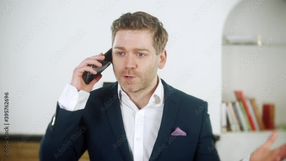 Handsome young businessman is shouting instructions on his phone. He is angry. The man is wearing a blue suit and is at home in his living room. Handsome young businessman is shouting instructions on his phone. He is angry. The man is wearing a blue suit and is at home in his living room.