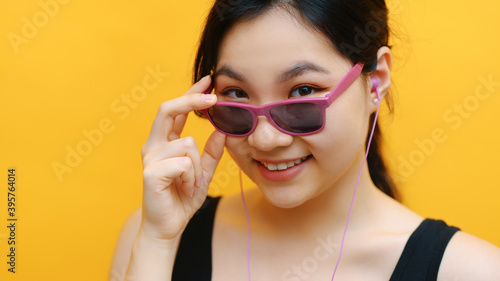 Portrait of young asian woman listening to the music on the headphones and looking over her pink sunglasess. Isolated on orange background. High quality photo