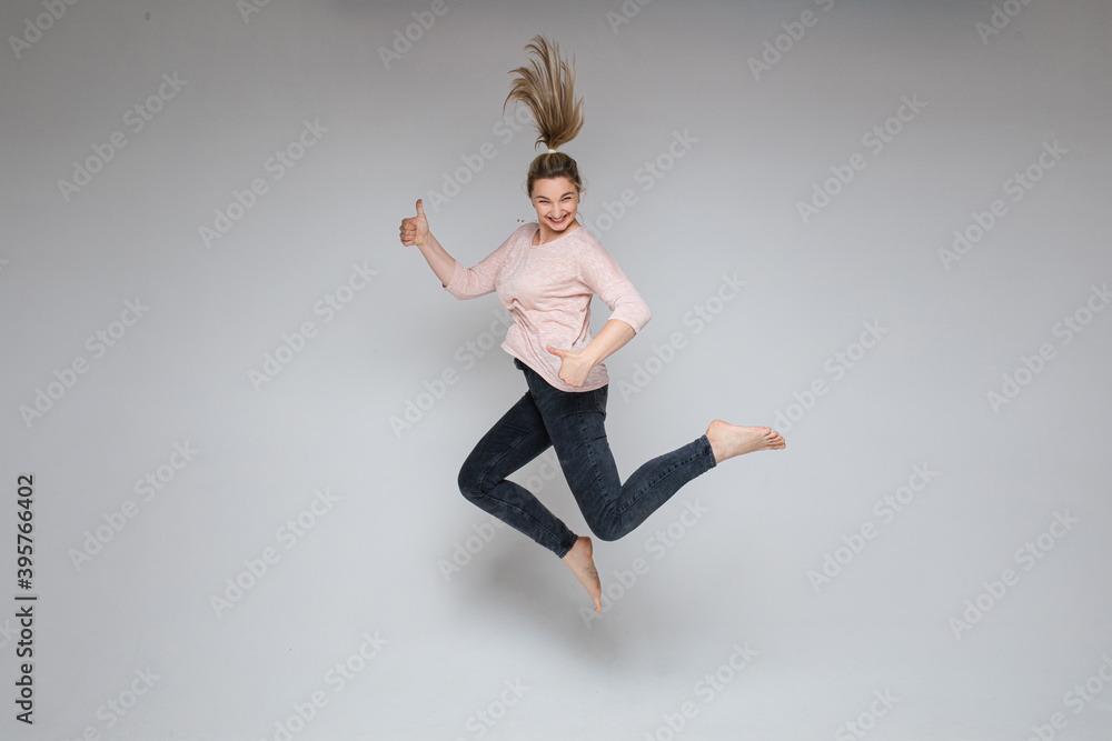 Stock photo of cheerful carefree blonde woman jumping in mid-air with thumbs up on white background. Jumping woman smiling at camera holding thumbs up.