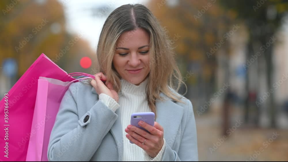 Young happy smiling shopaholic woman walking street and typing on smartphone. Lady smiling chatting and holding shopping bags. Young happy smiling shopaholic woman walking street and typing on smartphone. Lady smiling chatting and holding shopping bags.
