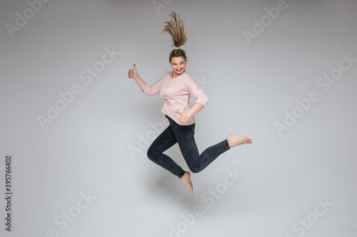 Fototapeta Naklejka Na Ścianę i Meble -  Stock photo of cheerful carefree blonde woman jumping in mid-air with thumbs up on white background. Jumping woman smiling at camera holding thumbs up.