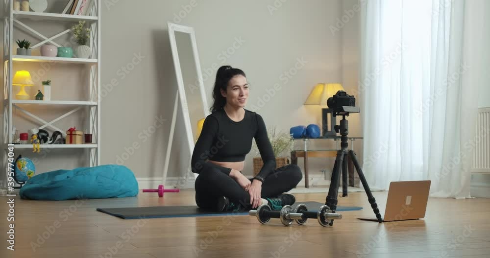 Young beautiful sport blogger recording video at home. Wide shot portrait of smiling confident Caucasian sportswoman sitting on exercise mat and talking. Sport and blogging. Cinema 4k ProRes HQ. Young beautiful sport blogger recording video at home. Wide shot portrait of smiling confident Caucasian sportswoman sitting on exercise mat and talking. Sport and blogging. Cinema 4k ProRes HQ.
