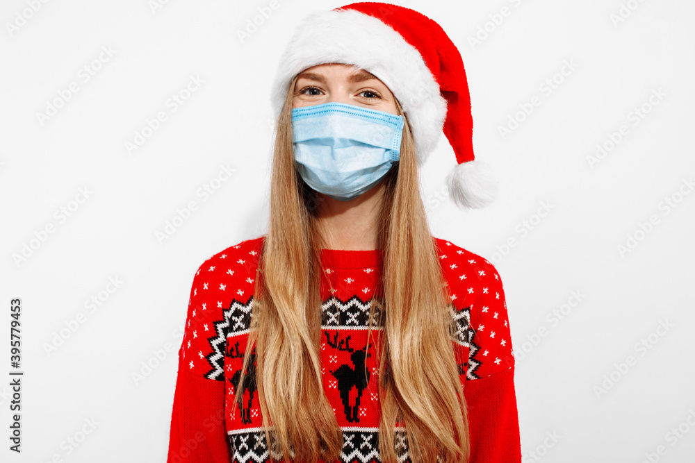 Young woman in a Christmas sweater and Santa Claus hat, in a medical protective mask on her face, from the covid-19 coronavirus, on a white background