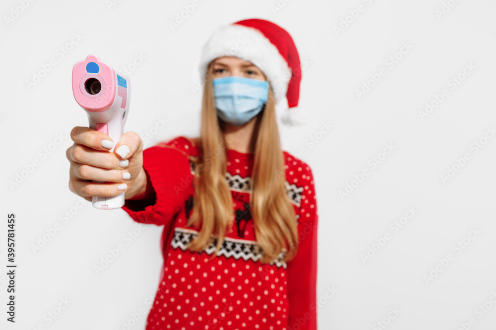 Young woman wearing a Christmas Santa Claus hat and a medical mask on her face, holding an infrared thermometer to check her body temperature, on a white background Young woman wearing a Christmas Santa Claus hat and a medical mask on her face, holding an infrared thermometer to check her body temperature, on a white background