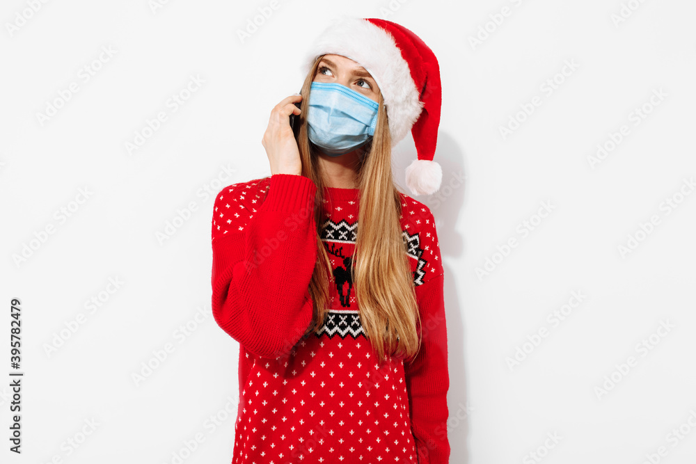 Young happy christmas woman, wearing a Santa Claus hat and a medical mask on her face, talking on the phone, holding a smartphone, on a white background Young happy christmas woman, wearing a Santa Claus hat and a medical mask on her face, talking on the phone, holding a smartphone, on a white background