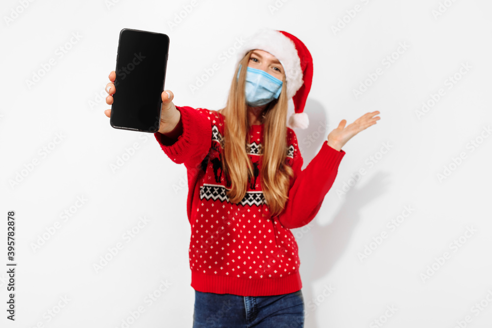Excited happy young woman wearing santa claus hat and medical protective mask, showing blank smartphone screen on white background Excited happy young woman wearing santa claus hat and medical protective mask, showing blank smartphone screen on white background