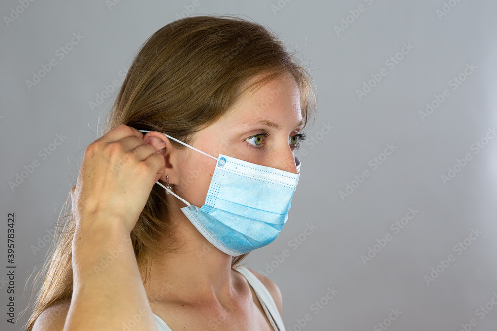 Young blond woman attaching surgical mask to her face. Adult person wearing respiratory protection and adjusting position of a earloop with her hand. Young blond woman attaching surgical mask to her face. Adult person wearing respiratory protection and adjusting position of a earloop with her hand.