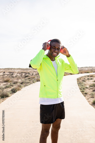 Vertical shot of a Young smiling black runner in a yellow rain jacket who enjoys listening to music on his cell phone before or after training. © menudencias