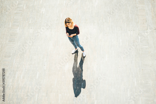 young blonde woman in casual clothes walks across empty paved city square looking at modern smartphone screen on sunny summer day upper view
