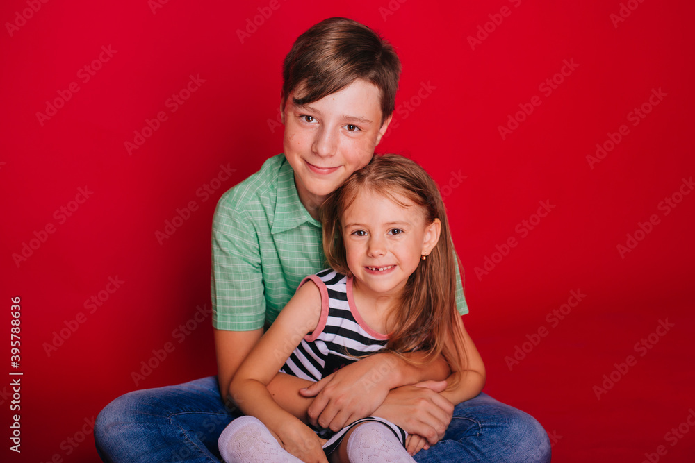 Brother and sister. Little girl in a striped dress with a boy in a green shirt on a red background. Brother hugs his sister. Brotherly love