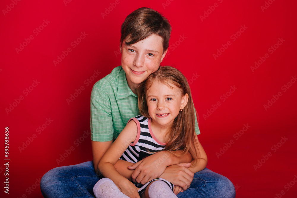 Brother and sister. Little girl in a striped dress with a boy in a green shirt on a red background. Brother hugs his sister. Brotherly love Brother and sister. Little girl in a striped dress with a boy in a green shirt on a red background. Brother hugs his sister. Brotherly love