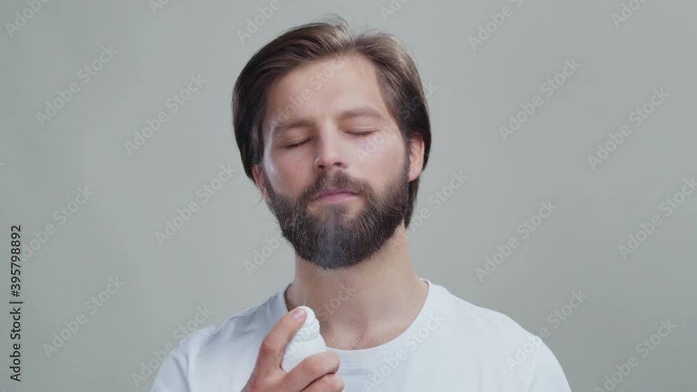 Handsome caucasian man with beard spraying moisturizing mist spray on face advertising at grey background. Person's beauty concept. Skincare. Handsome caucasian man with beard spraying moisturizing mist spray on face advertising at grey background. Person's beauty concept. Skincare.