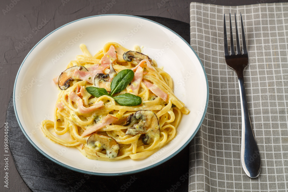 a plate of pasta with ham and mushrooms in a creamy sauce with basil on a black round stand with a fork on a napkin.