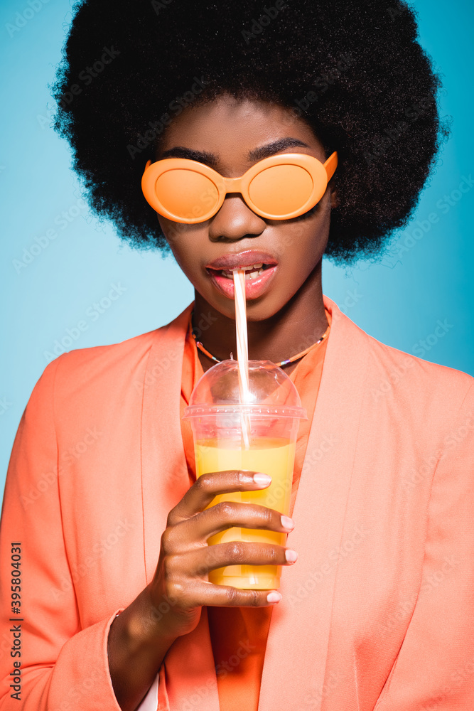 african american woman in stylish outfit with orange juice isolated on blue background african american woman in stylish outfit with orange juice isolated on blue background