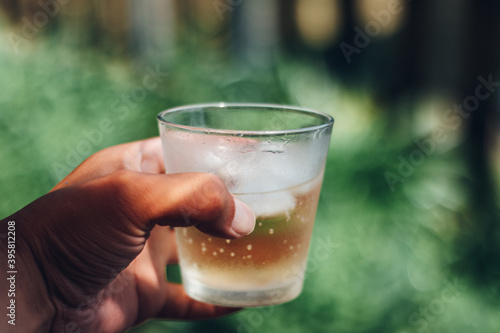 Holding a drink and ice in the glass against yard green background in summer