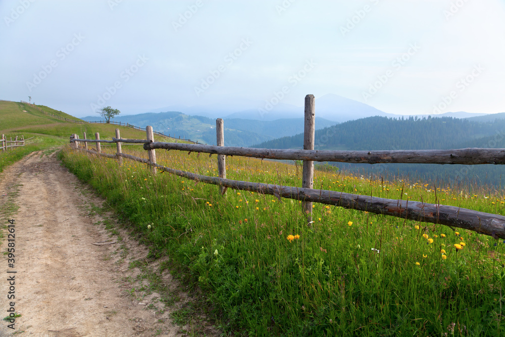 Mountain landscape, road with a wooden fence along a bright meadow, silhouettes of mountains on the horizon, grey sky. Ukraine, Carpathians.