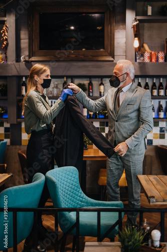 Confident senior businessman standing in exclusive restaurant. He takes off his coat while the waitress helps him. They both wearing protective face masks against virus infection. © Dusko
