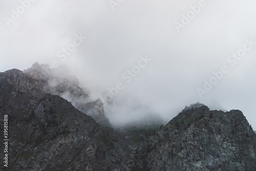 Dramatic bleak fog among giant rocky mountains. Ghostly atmospheric view to big cliff in cloudy sky. Low clouds and beautiful rockies. Minimalist scenery in mysterious place at early foggy morning.