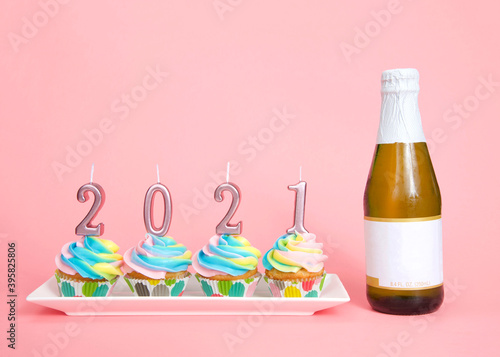 Colorful rainbow frosted vanilla cupcakes sitting in a row on a white, rectagular porcelain plate with 2021 candles on top next to a small bottle of wine, blank label. Pink background photo