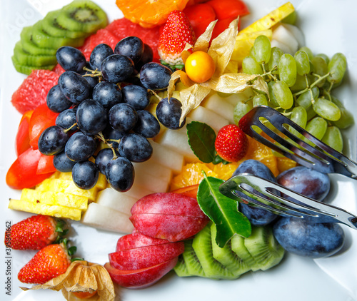 Assorted sliced fruit  black and white grapes  kiwi  prunes  pineapple  strawberries and physalis are on a platter. The view from the top.